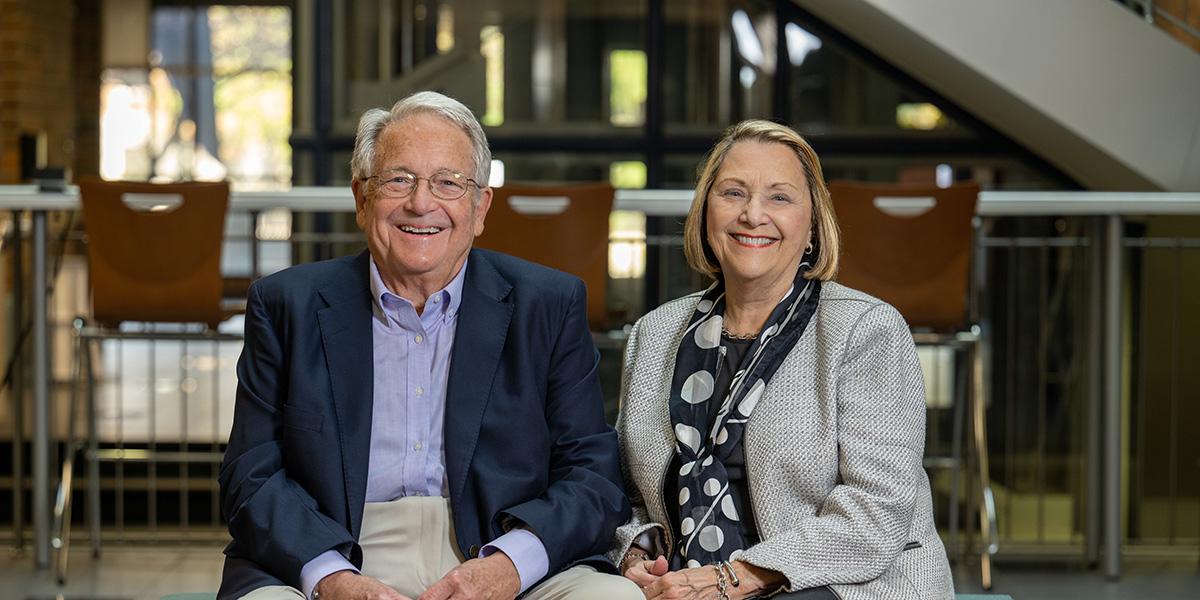 A smiling couple, Gloria and Steve Bailey, seated in the Scott College of Business. Steve is wearing a light blue shirt, a dark blue jacket, and beige pants. To his right, Gloria is wearing a business casual white jacket with dark blouse and pants and a black scarf with large white circles on it.