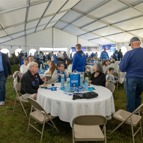 A large group of Indiana State University alumni and friends gather under a large tent for tailgating, sitting around tables with drinks and snacks, wearing ISU gear.